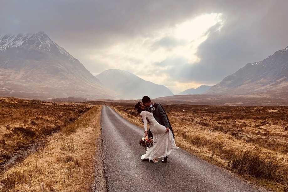 Groom dips bride holding bouquet in kiss with snow covered mountain tops in background